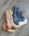 Tan cowboy boots next to a denim vest with lace trim, all lying on a gray wooden floor.