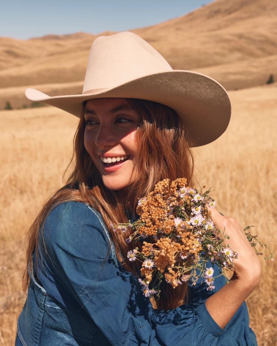 A person wearing a cowboy hat smiles while holding a bouquet of wildflowers in a dry, grassy field under a clear blue sky.