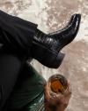 A person in elegant attire with a crocodile leather shoe, resting their foot on a knee, holding a glass of amber liquid.