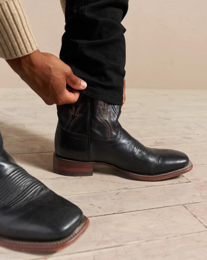 A person wearing black leather boots is adjusting the cuff of their black pants.