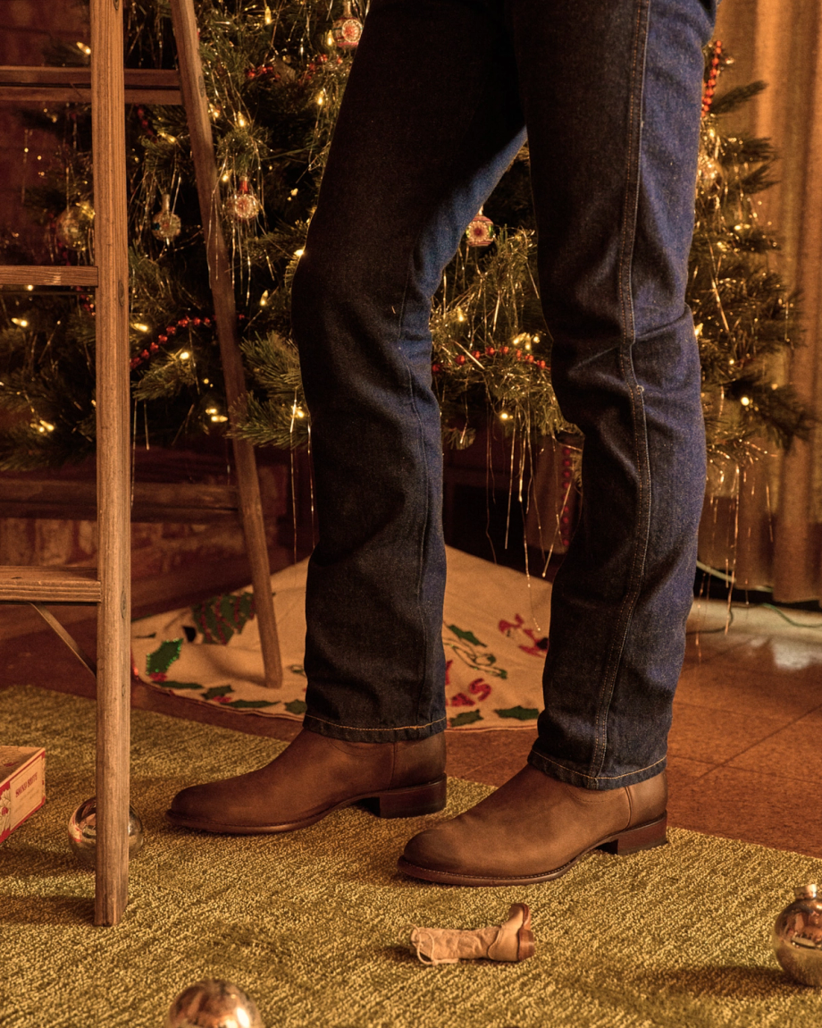 The Earl in Cafe Goat. Person in jeans and brown boots stands near a wooden ladder and decorated Christmas tree, with ornaments, a small nativity figure on a green rug, and a goat figurine reminiscent of the festive charm at Cafe Goat.