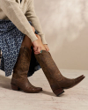 Person sitting, wearing a patterned blue skirt and beige sweater, adjusting tall brown cowboy boots.