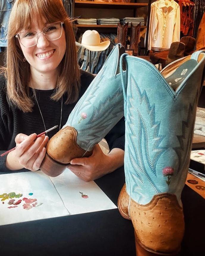 A woman smiles while hand-painting designs on a pair of light blue cowboy boots at a table with paint and brushes.