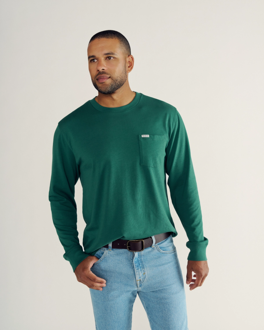 A man wearing a green long-sleeve shirt and blue jeans stands against a plain light background, looking slightly to the side.