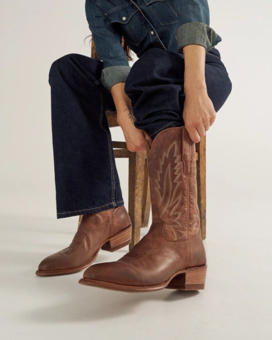 Person in denim outfit sits on a stool, pulling on a pair of brown leather cowboy boots with decorative stitching.
