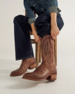 Person in denim outfit sits on a stool, pulling on a pair of brown leather cowboy boots with decorative stitching.