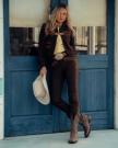 A woman in a black and gold western outfit holds a white cowboy hat, standing in front of a blue wooden door and window at The Tess Goat cafe.