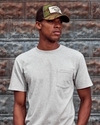 Man wearing the standard issue pocket tee in heather grey against a rustic wall