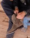 A person is sitting on a bench and zipping up a pair of brown suede cowboy boots. The individual is dressed in dark jeans and a jacket. The setting appears to be rustic with a wooden floor.
