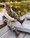 A young person in camouflage pants and boots sits on the edge of a docked metal boat by the water, smiling and holding one boot.
