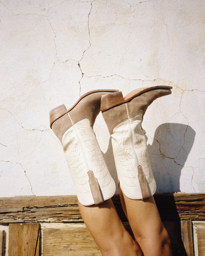 Person's legs against a cracked wall, wearing beige cowboy boots with decorative stitching.