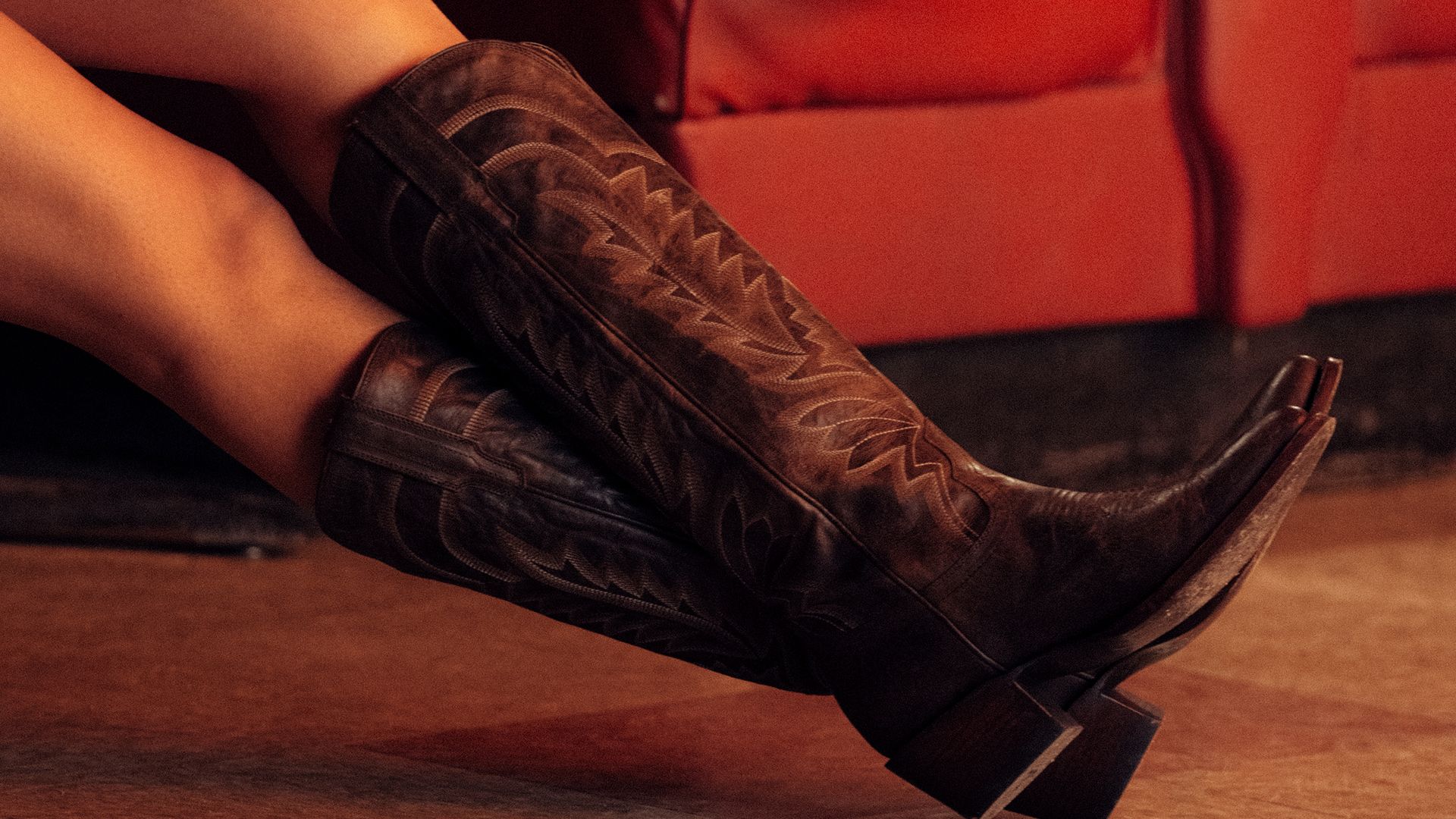 A person wearing dark brown cowboy boots with decorative stitching, resting their legs on a red seat.