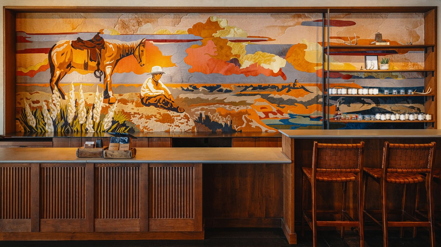 A mural of a cowboy and horse in a western landscape decorates the wall behind a wooden counter and shelves in a rustic-style café with two bar stools.
