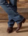 A person in blue jeans and brown cowboy boots stands with legs crossed on a concrete floor, with a wooden wall in the background.