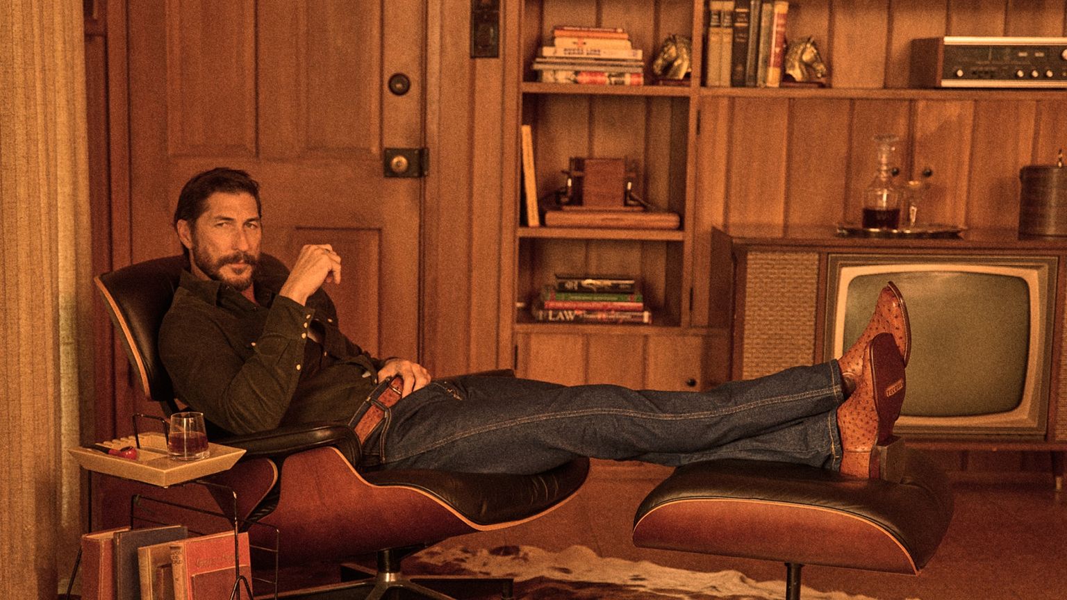 A man with a beard lounges on a chair with his legs up on an ottoman in a wood-paneled room, next to a vintage TV, bookshelves, and a drink on a side table.