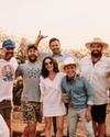 A group of eight people outdoors, some wearing hats, smiling and posing together with drinks in hand. Trees and a sunset are in the background.