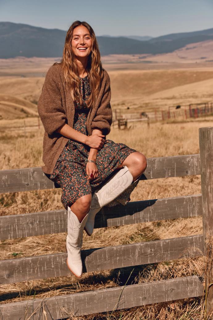 A woman in a floral dress and boots sits on a fence in a wide-open field with mountains in the background.