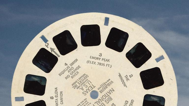 A close-up of a white vintage View-Master reel labeled "BIG BEND NATIONAL PARK TEXAS" held up against a blue sky.