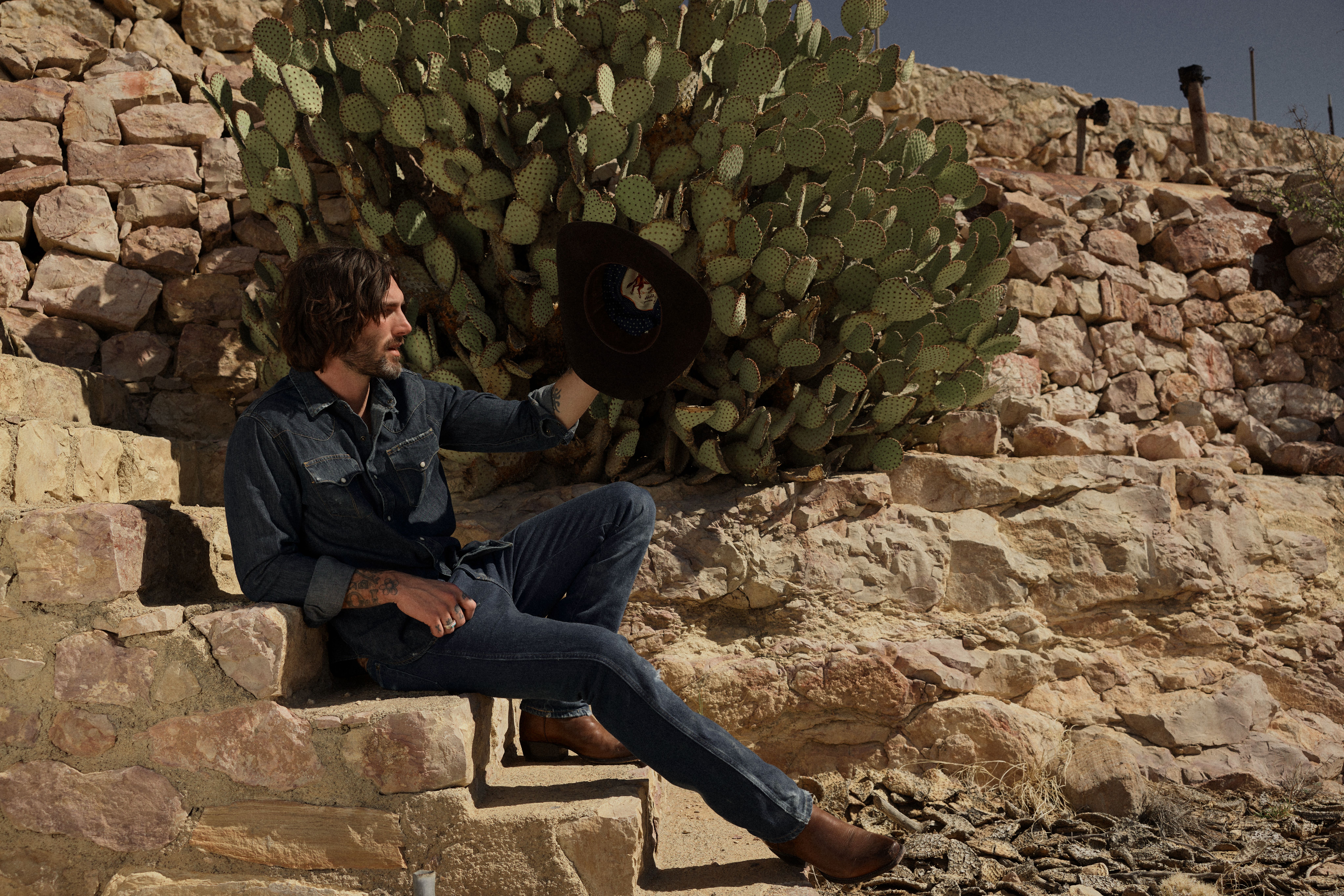 A man in denim clothing sits on stone steps beside a cactus, holding a hat in one hand, with a stone wall in the background under a clear sky.