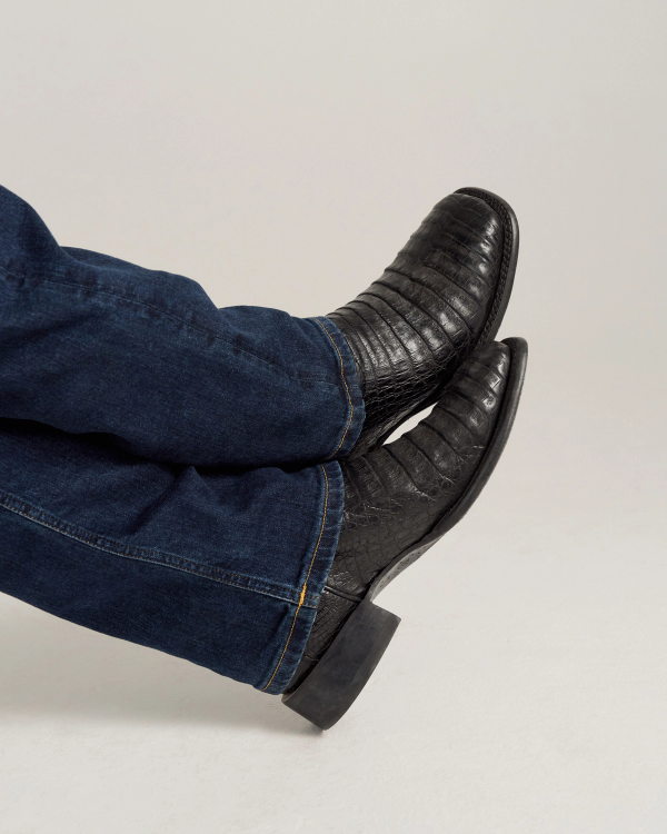 The William in Midnight Caiman. A person wearing dark blue jeans and black textured Midnight Caiman boots is sitting with their legs crossed against a plain background.