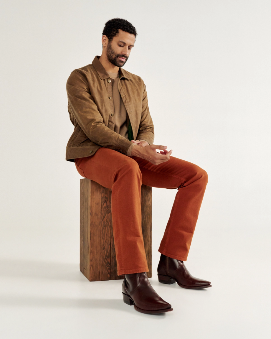 Man seated on a wooden block wearing a brown jacket, beige shirt, orange pants, and dark boots, looking at a small object in his hands against a plain background.