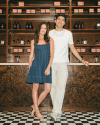 A woman in a blue dress and a man in a white outfit stand side by side in front of a wooden shelf with various jars and boxes, with the man leaning on the counter.