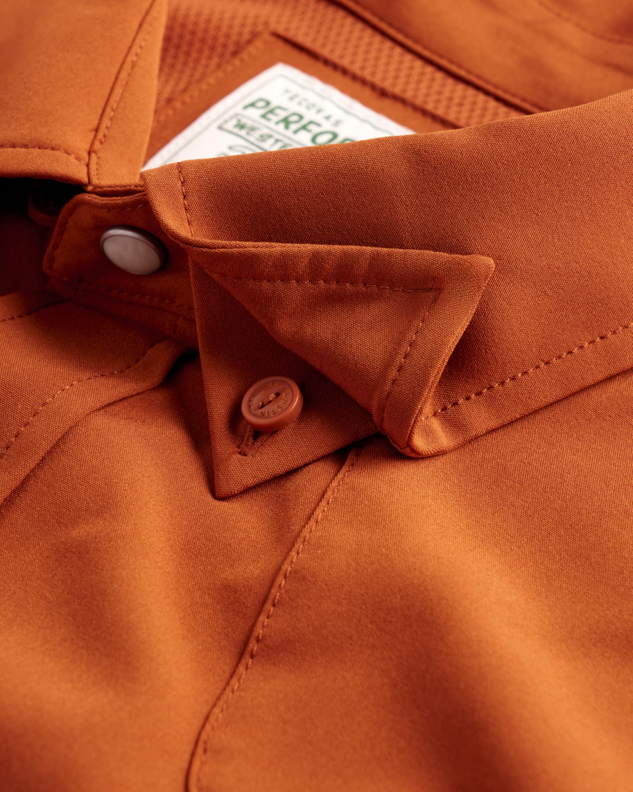Close-up of a rust-colored collared shirt, showing the collar, a white snap button, a brown button, and part of a label inside the neckline.