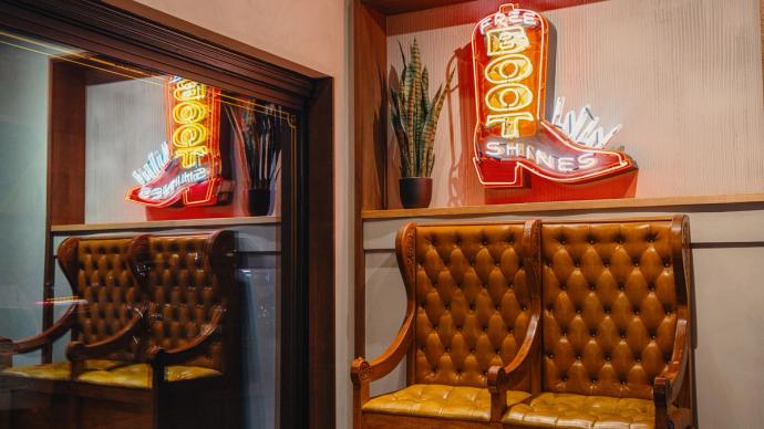 A room with leather chairs and a neon sign shaped like a boot that reads "FREE BOOT SHINES." The sign is illuminated and there are plants on the shelf above the chairs.