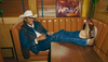 Person in a cowboy hat and denim outfit lounges on a brown leather booth seat with feet up, holding a drink.