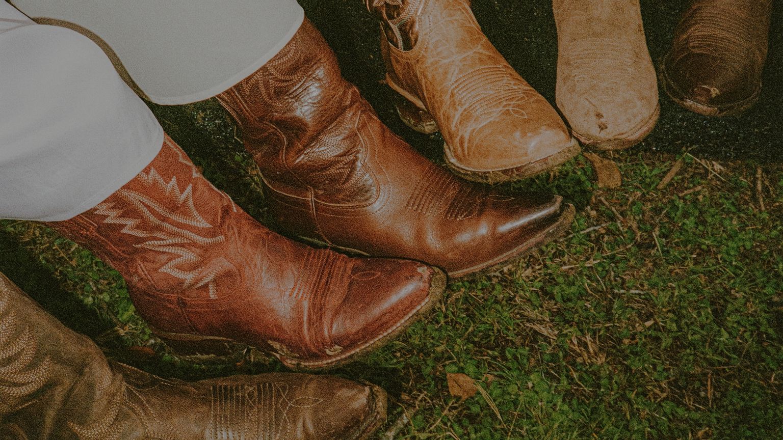 Five pairs of cowboy boots in varying shades of brown and tan are arranged in a circle on grass, some worn and some newer.