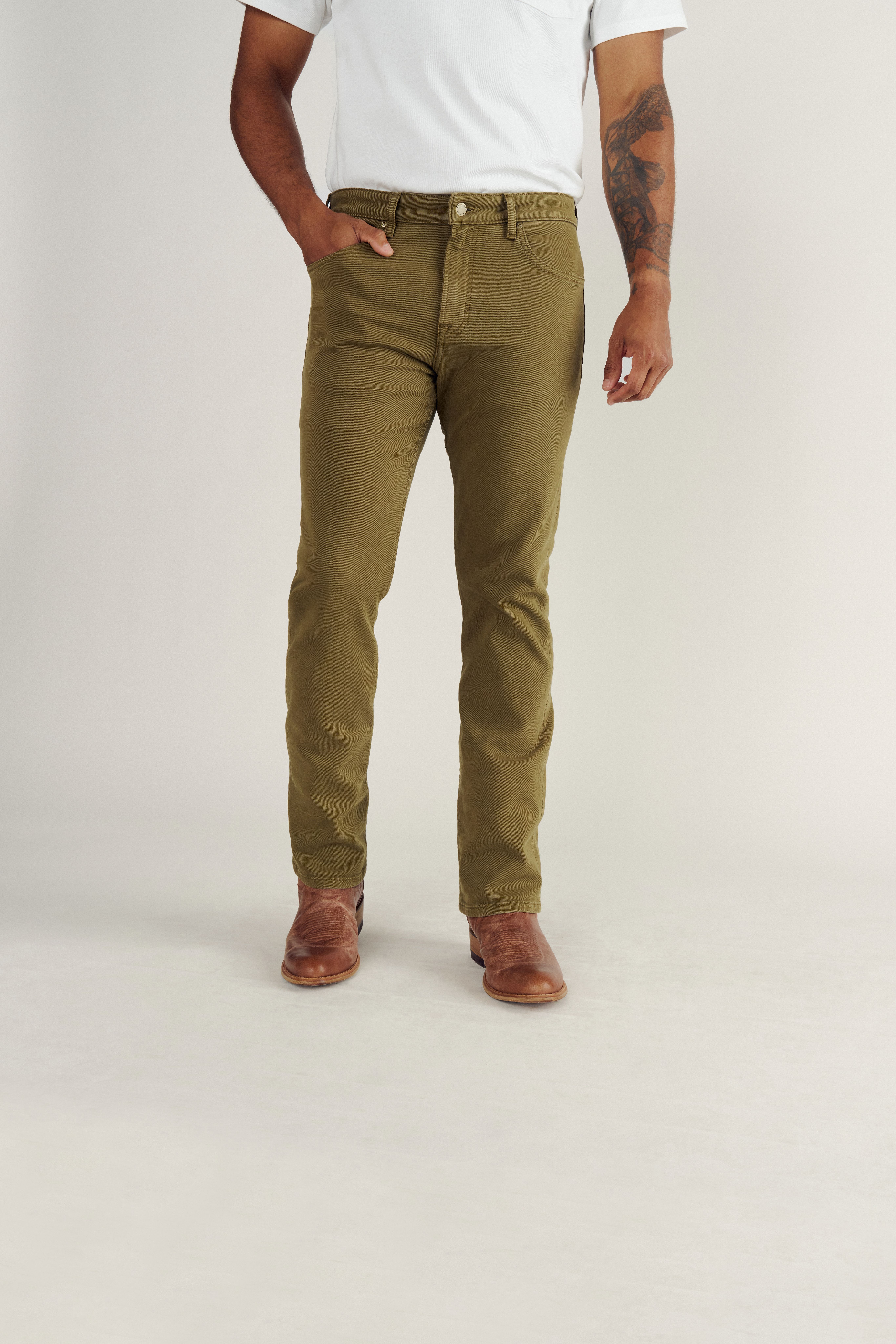 A person wearing a white t-shirt, olive green pants, and brown shoes stands with one hand in their pocket against a plain background.