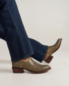 A person wearing dark blue jeans and brown textured leather ankle boots with a low wooden heel stands on a light surface.