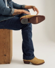 A person wearing blue jeans and tan cowboy boots sits on a wooden bench with one leg crossed, showing the sole marked "TECVAS.