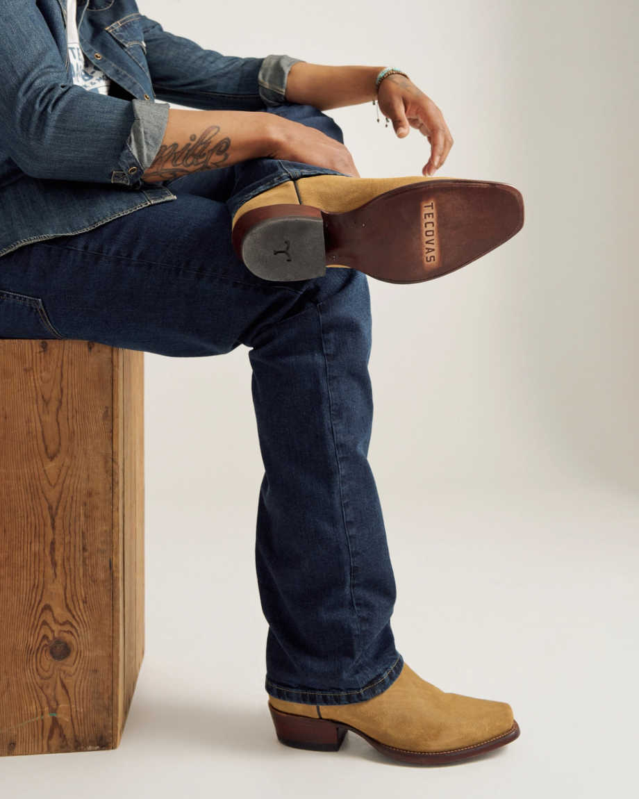 A person wearing blue jeans and tan cowboy boots sits on a wooden bench with one leg crossed, showing the sole marked "TECVAS.