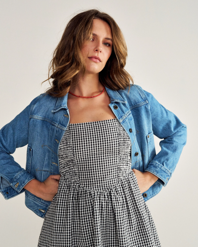 A woman stands with her hands on her hips, wearing a blue denim jacket over a black and white checkered dress, against a plain background.