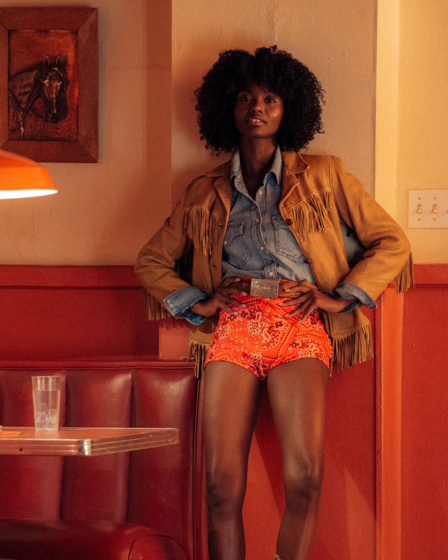 A person with curly hair stands in a retro diner, wearing a fringed jacket, denim shirt, and red patterned shorts, with hands on hips.