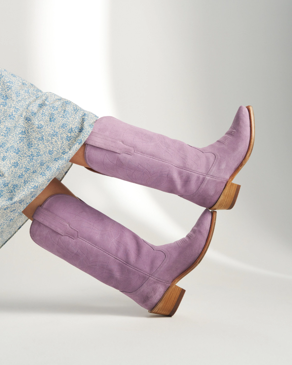 A person wearing a light blue floral dress and The Annie Suede knee-high lavender boots with wooden heels, legs extended and crossed at the ankles.