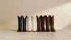 A row of seven cowboy boots in various colors, including black, white, and brown, lined up against a beige wall on a wooden floor.