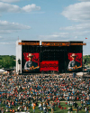 Large outdoor concert, daytime. A stage displays a musician on screen with "Barley Cook Red" text. Numerous attendees seated on grass, enjoying the performance under a partly cloudy sky.