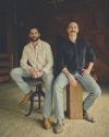 Two men with mustaches sitting on stools wearing jeans and boots