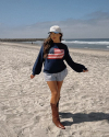 Person standing on a beach wearing a navy sweater with an American flag, striped shorts, a white cap, and brown boots. The ocean and a rocky jetty are visible in the background.