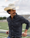 A person with a mustache wearing a cowboy hat and dark shirt stands by a railing in front of a sports field.