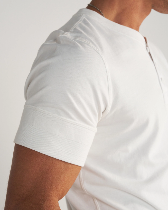 A person wearing a white short-sleeve t-shirt, blue jeans, and a cream-colored cowboy hat stands facing away from the camera against a plain background.