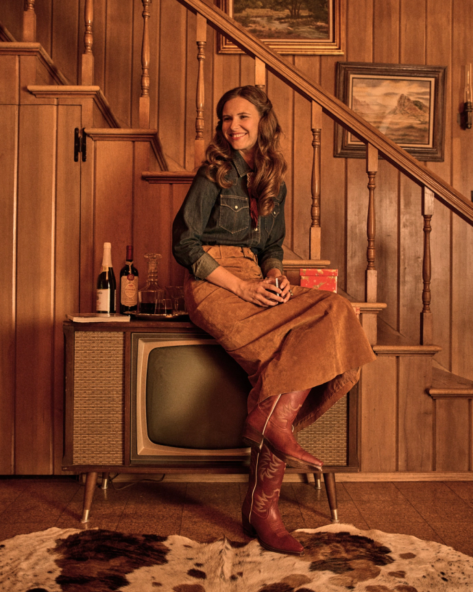 The Annie in Scarlet Cowhide. A woman in a denim shirt, brown skirt, and scarlet cowboy boots sits on a vintage TV in a wood-paneled room with drinks, paintings, and a scarlet cowhide rug.