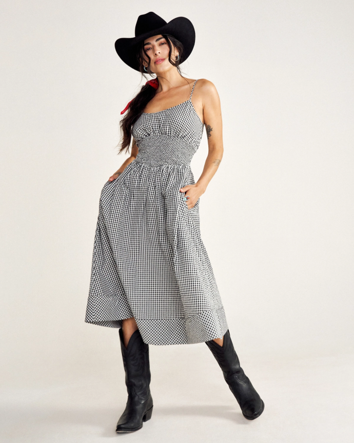 A person wearing a black hat, checkered dress, and black cowboy boots poses against a plain background.