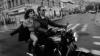 A man rides a motorcycle with a woman sitting behind him on a city street. The image is in black and white and captures motion blur in the background.