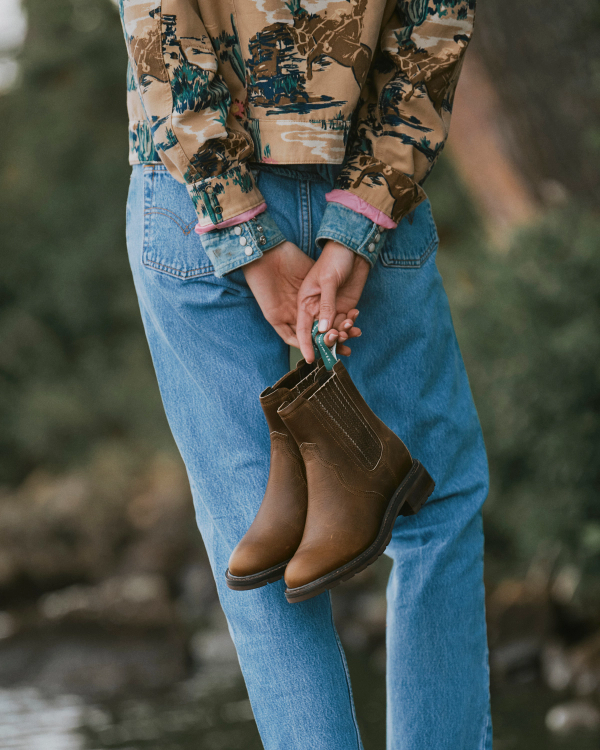 Person in blue jeans and patterned shirt stands outdoors, holding brown leather boots behind their back, with greenery blurred in the background—channeling the relaxed spirit of a weekend at Cafe The Chelsea Cowhide.