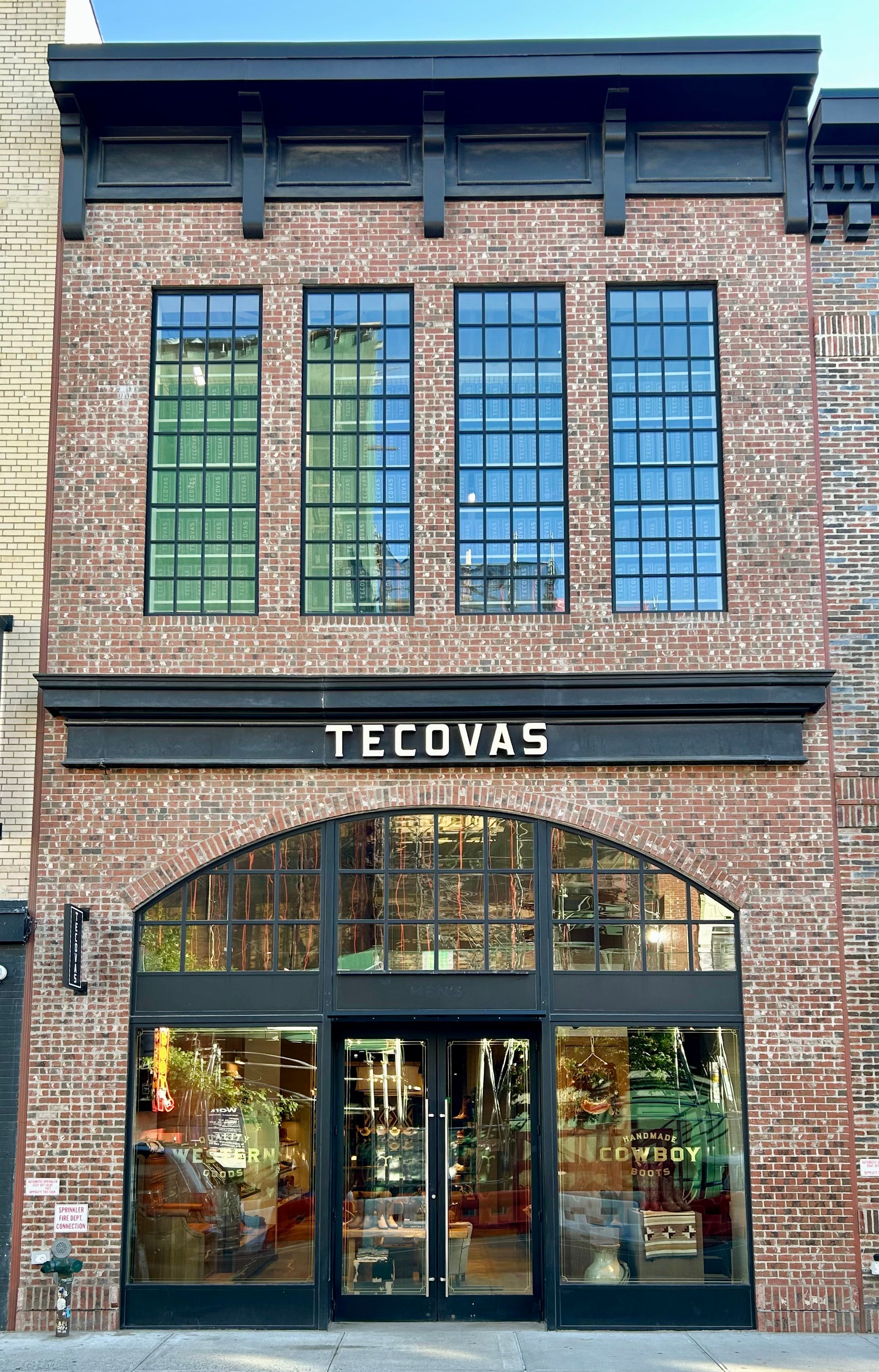 Exterior view of a Tecovas store with large brick facade, tall grid windows, and glass entrance doors displaying boots inside.