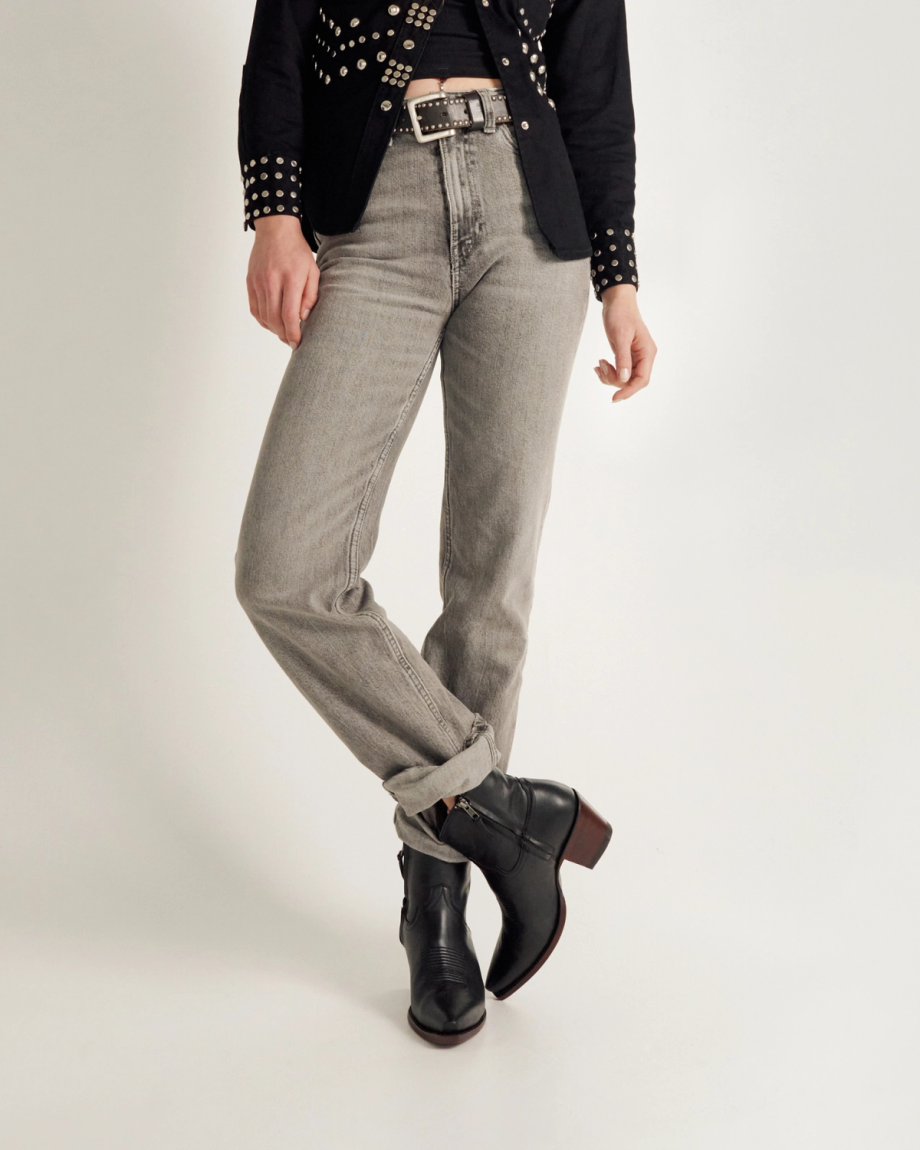 Person wearing light gray jeans, black ankle boots, a black belt with silver details, and a black studded jacket, standing against a plain white background.