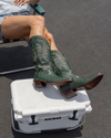 Person sitting on a folding chair, wearing denim shorts and green cowboy boots, with their feet resting on a white cooler.